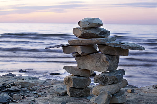 An inuksuk on a rocky shore against the sunset lit sky and waters of Georgian Bay in Craigleith Provincial Park near Collingwood, Ontario.